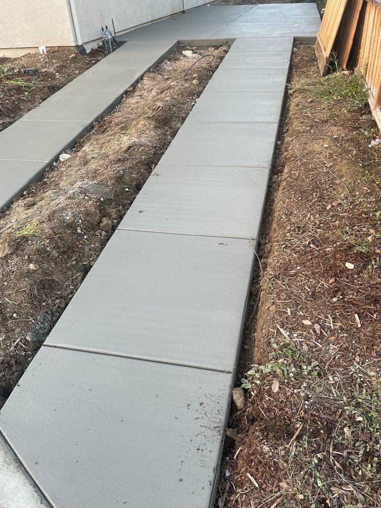 Concrete walkway beside a building, with a long narrow dirt strip in the middle and grass along the sides.
