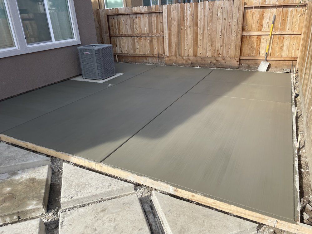 Freshly poured concrete patio beside a house and wooden fence, with an air conditioner unit nearby