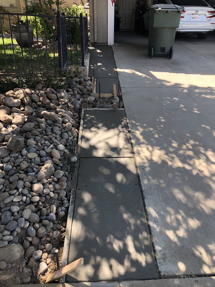 Broken sidewalk slabs beside a rock bed and driveway, with a trash bin in the background.