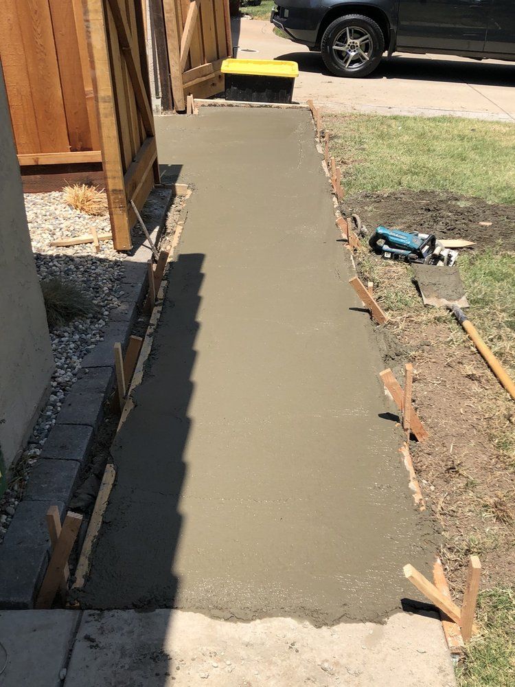 Freshly poured concrete walkway beside a wooden fence, leading to a driveway and parked SUV.