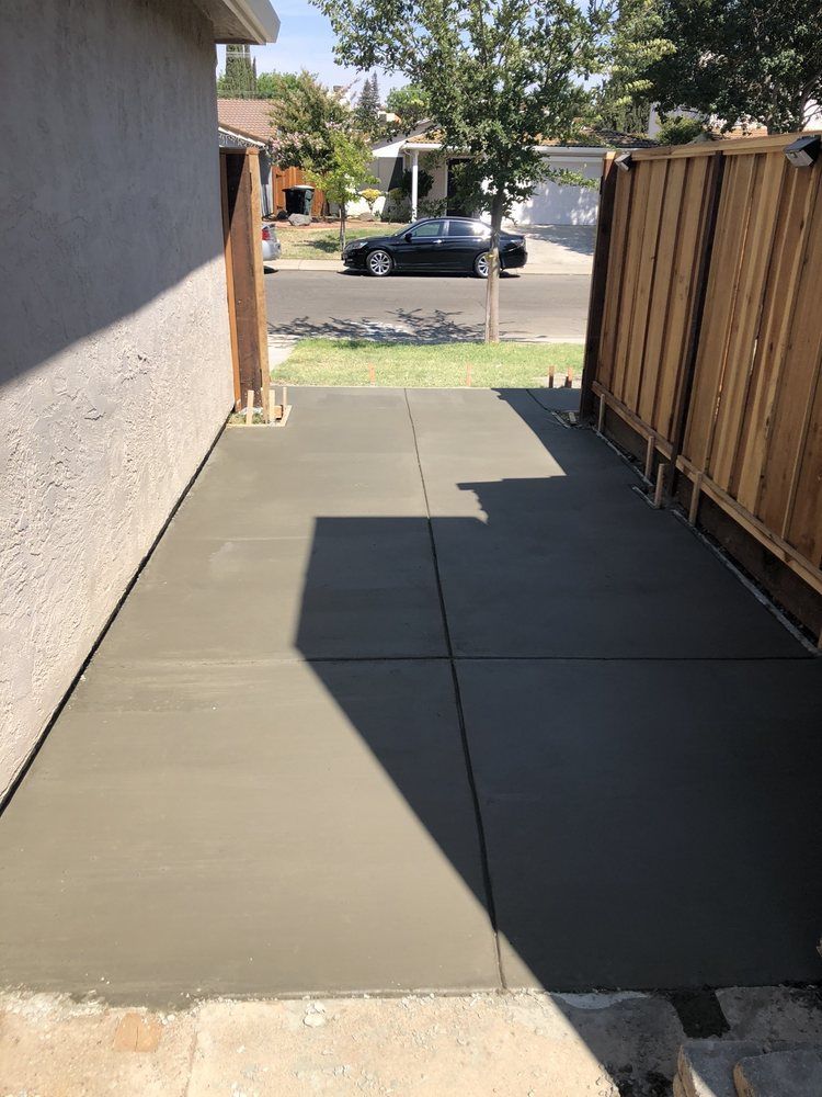 Freshly poured concrete walkway beside a house, bordered by a wooden fence and leading to a street.
