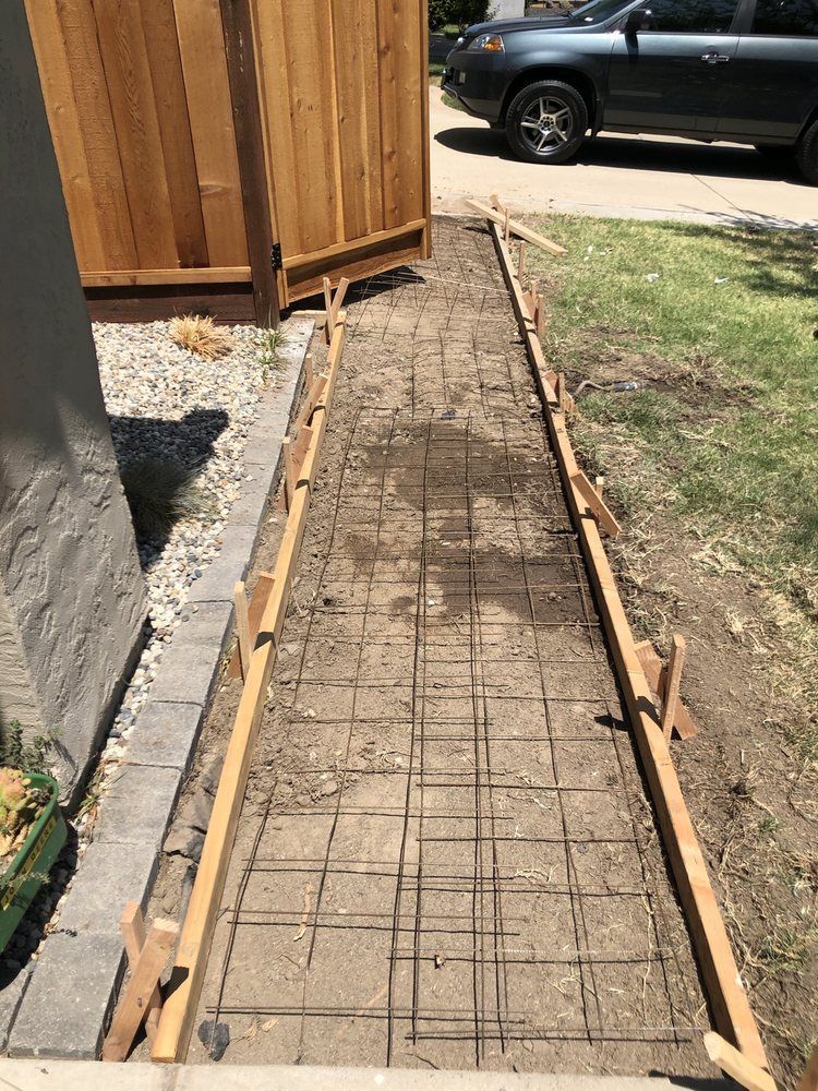 Fresh concrete walkway form with rebar beside a wooden fence and gravel driveway.