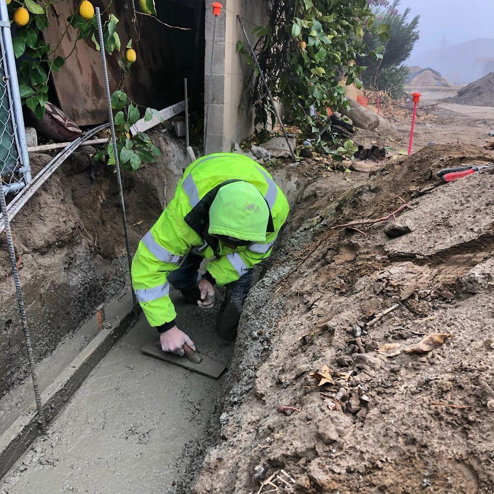 Worker in neon green jacket smoothing wet concrete in a trench beside muddy construction site.