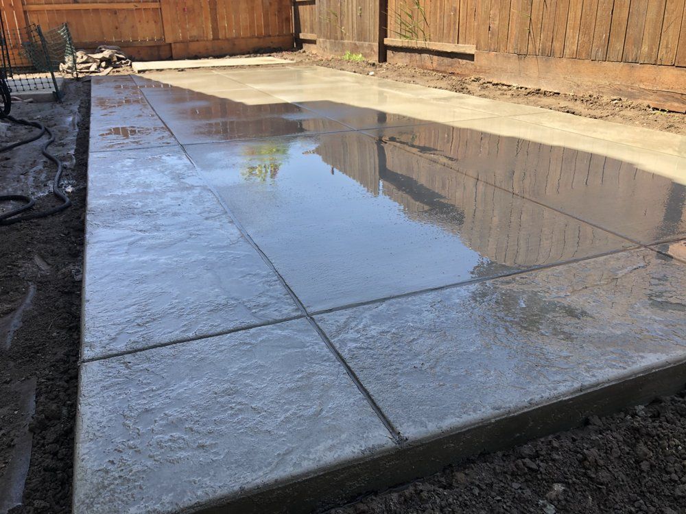 Wet concrete patio beside a wooden fence, reflecting light and the sky.