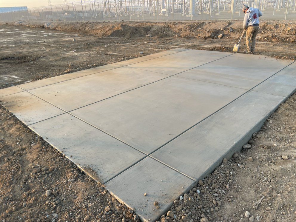 Concrete slab foundation in a dirt lot, with a worker standing nearby under construction.