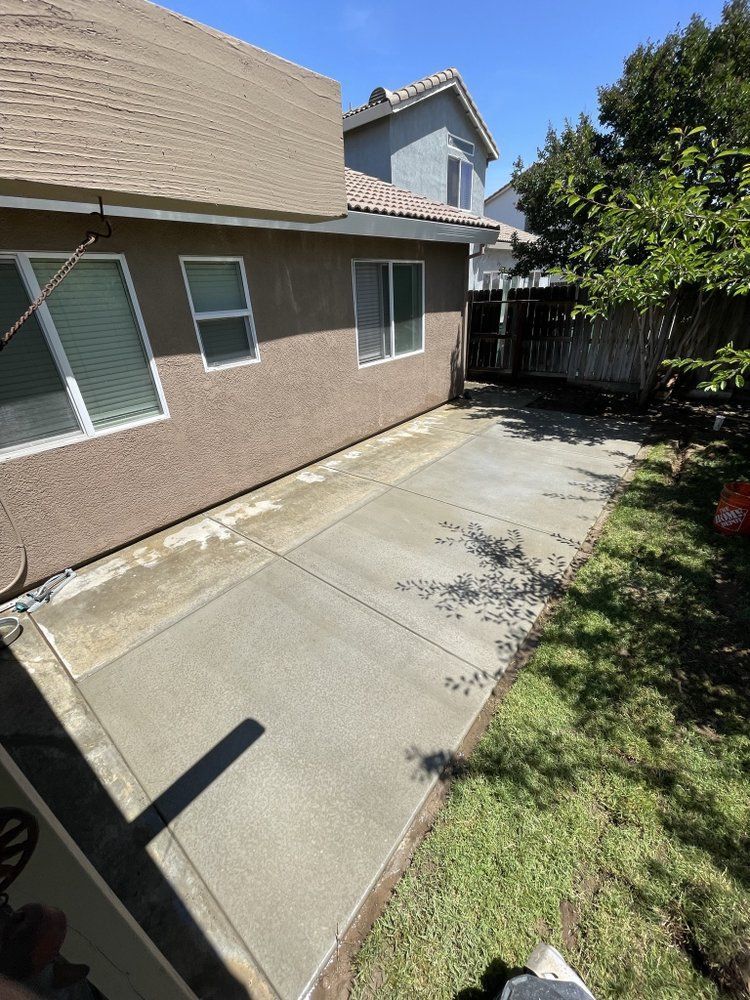 Side yard concrete patio beside a house, with grass border, fence, and sunlight.