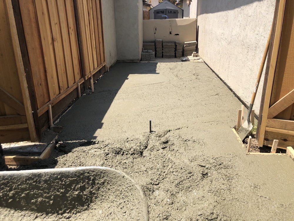 Narrow concrete walkway under construction between wooden fences, with fresh concrete and tools visible.