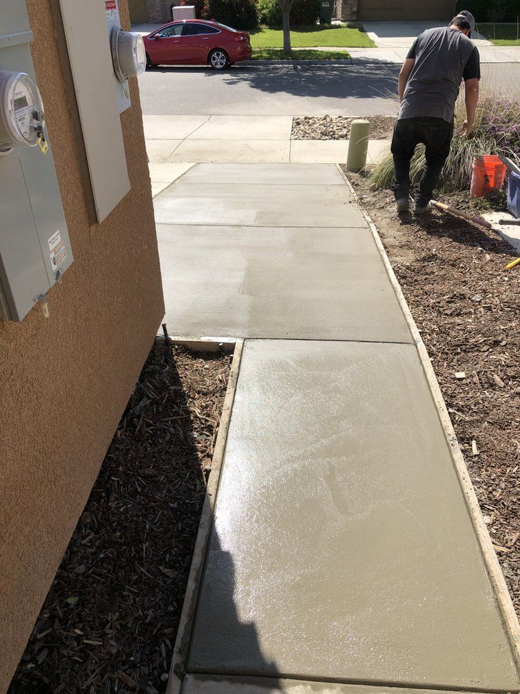 Fresh concrete sidewalk beside a house, with a person working near the edge and a red car in the driveway.