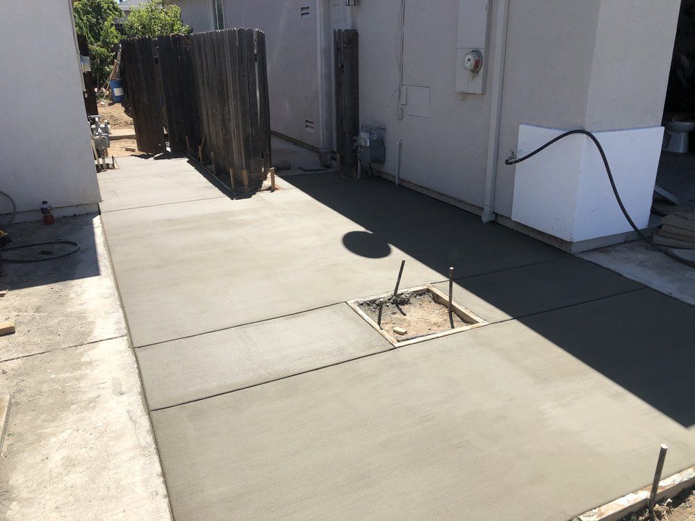 Concrete walkway beside a white building, with a small planted tree and dark fence shadows.