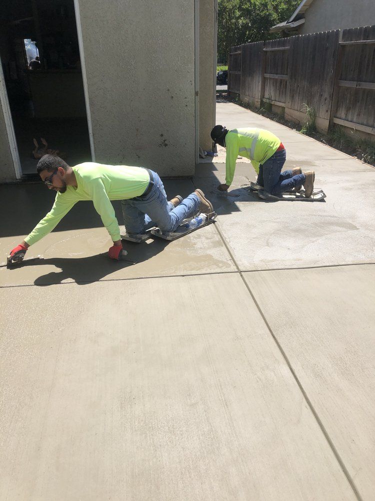 Two workers in neon safety shirts kneel and stretch concrete on a sunlit sidewalk.