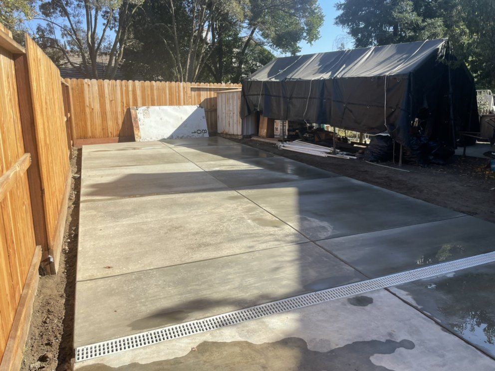 Empty concrete backyard with wooden fence, drainage grate, and a black covered trailer or shed on the right.