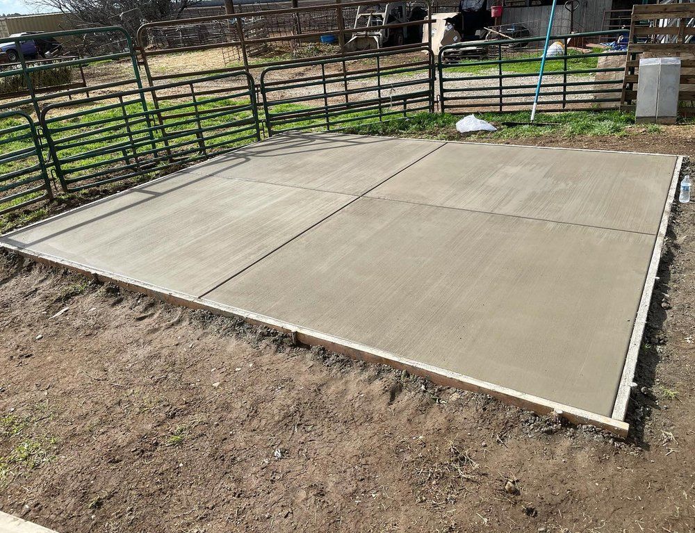 Freshly poured concrete slab in a fenced yard, surrounded by dirt and grass.