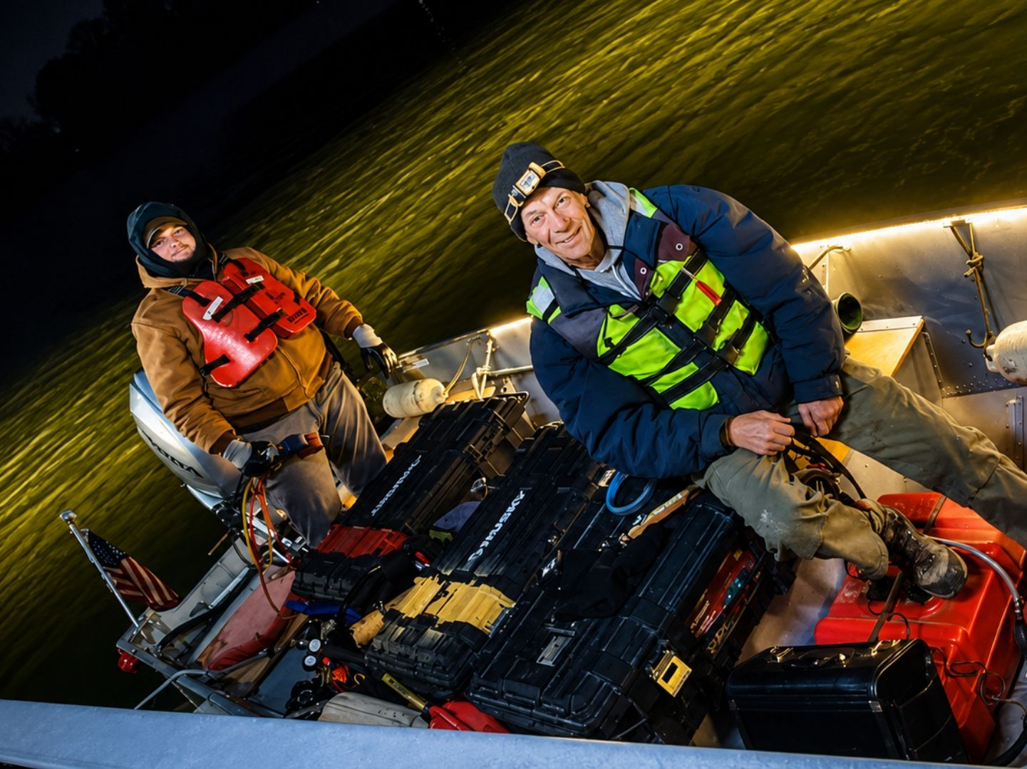 Two bundled-up people riding a snowy nighttime sled, lit by bright headlamps.