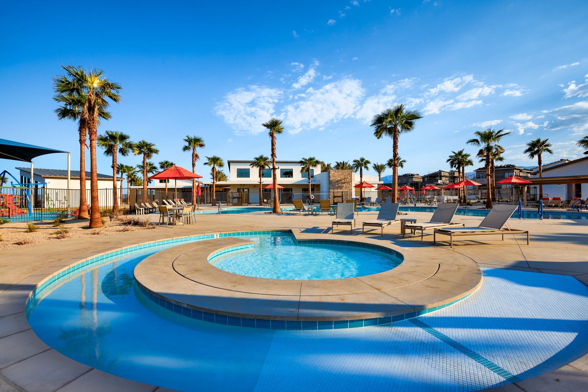 Swimming pool resort with palm trees, sunny sky, and buildings in the background.