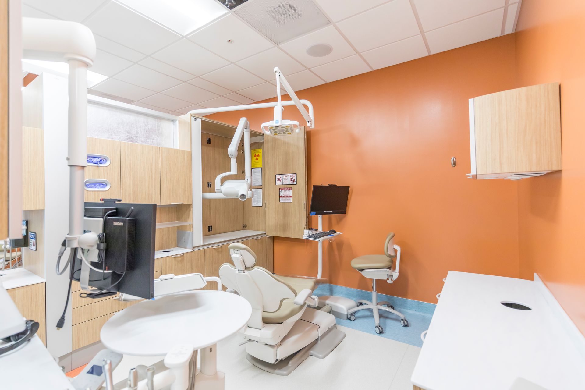 A dental examination room with a patient chair, dental equipment, a computer monitor, and light orange walls.