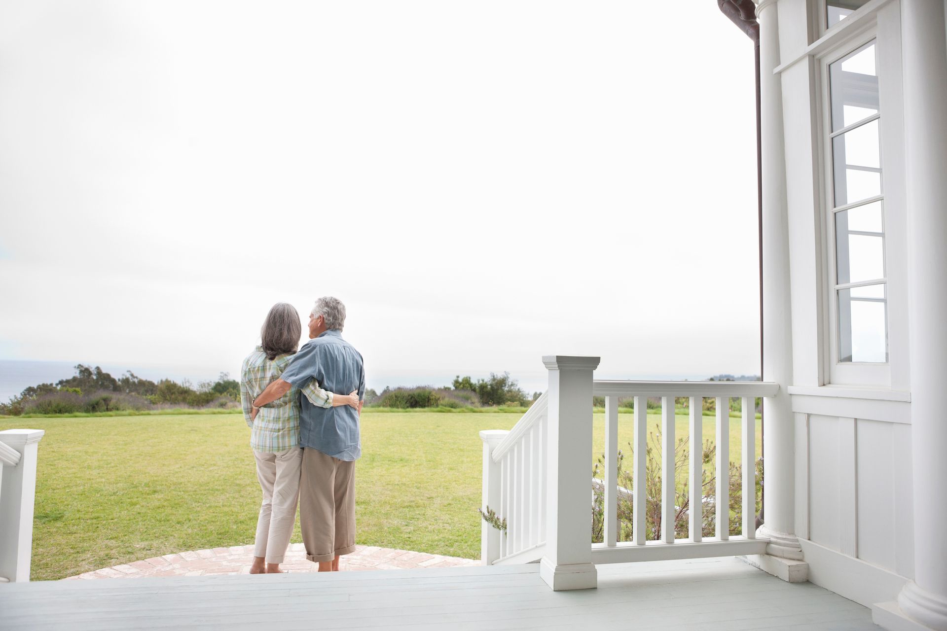 Couple with arms around each other, standing on a porch overlooking a green field and cloudy sky.