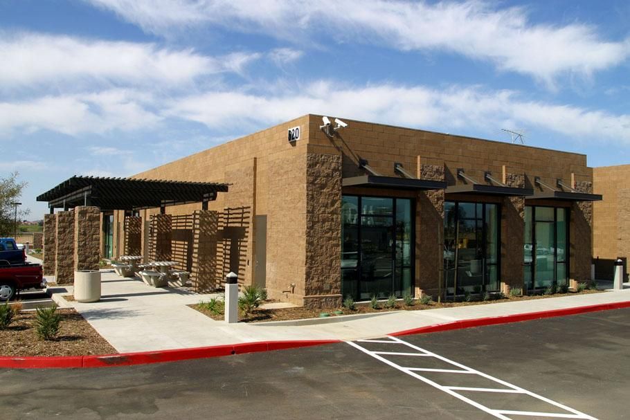 A modern building with an outdoor seating area under a pergola. Beige exterior, large windows, red curb.