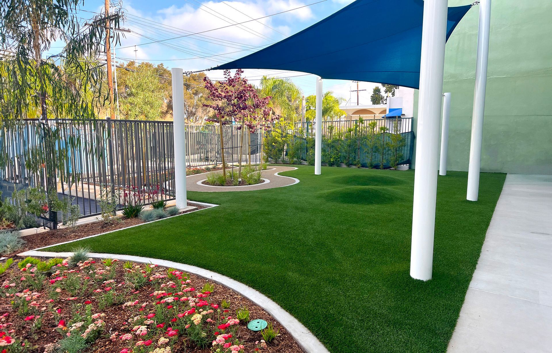 Outdoor play area with artificial turf, a shade sail, and garden beds.