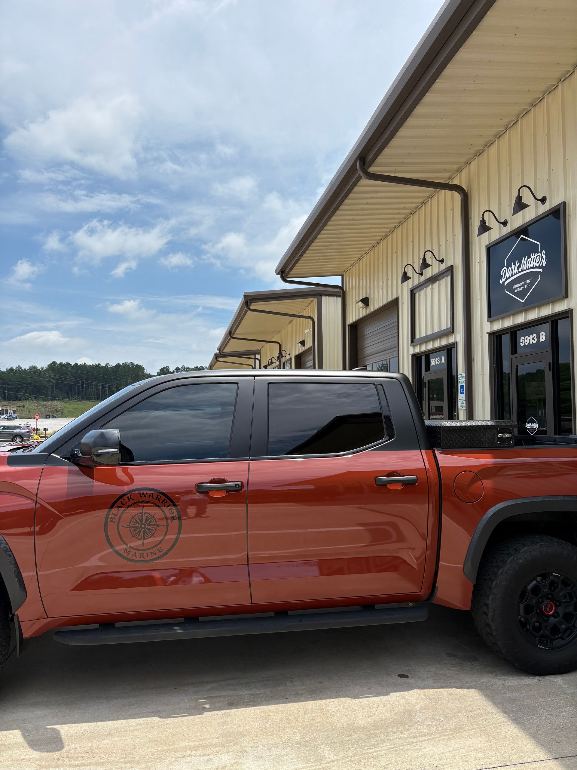 Rust-colored pickup truck parked outside a tan building with black signs. Overcast sky.