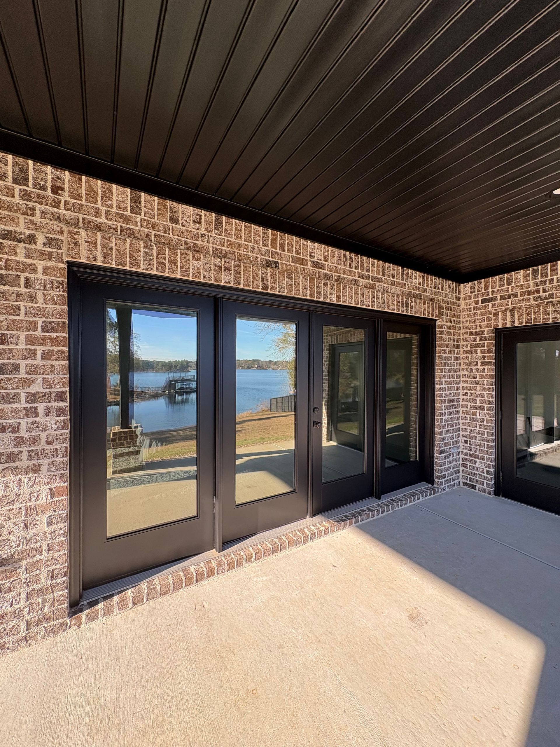 Exterior brick wall with black-framed glass doors overlooking a lake. A concrete patio and dark ceiling are also visible.