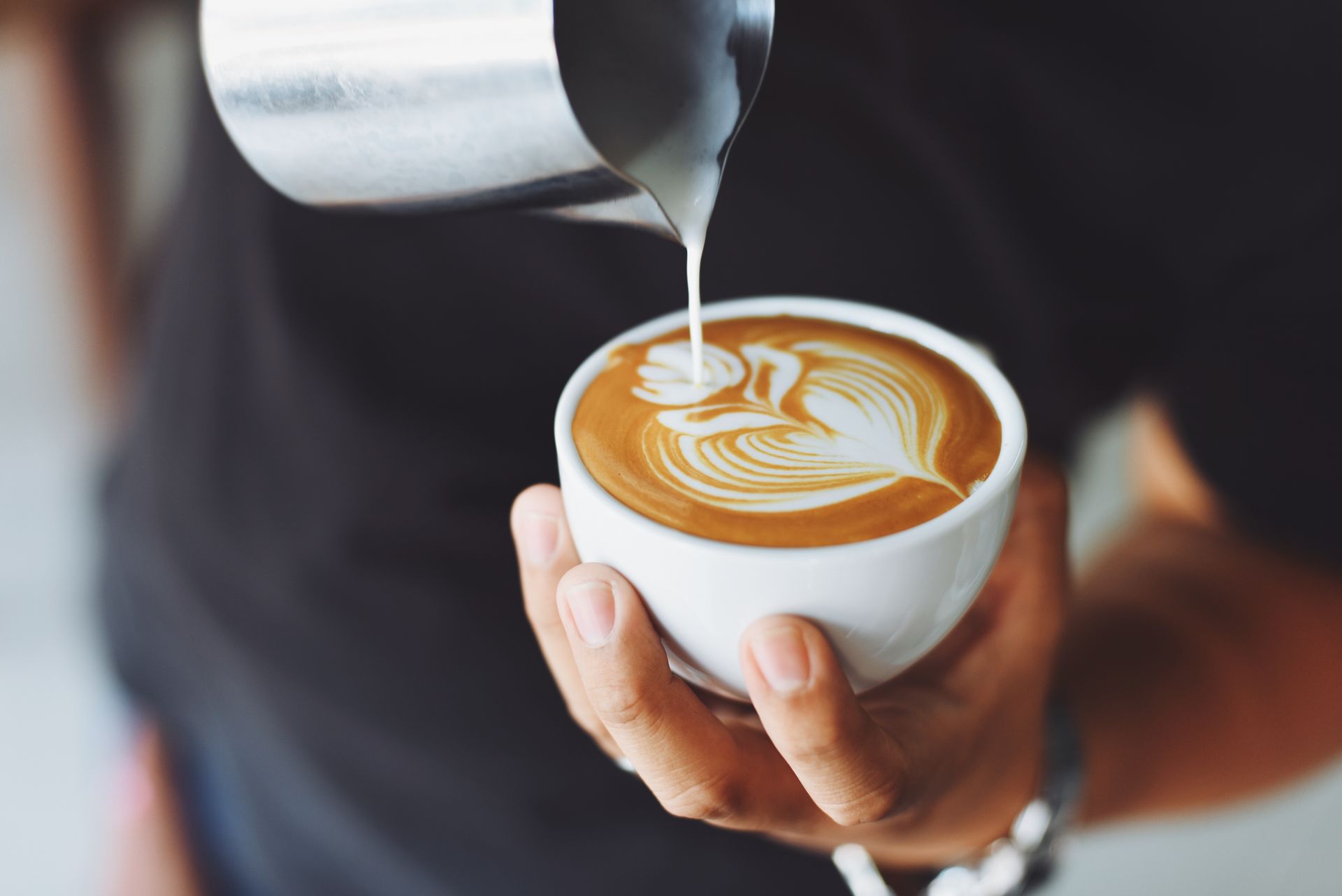 A barista pours steamed milk into a cup of coffee, creating a intricate rosetta latte art design.