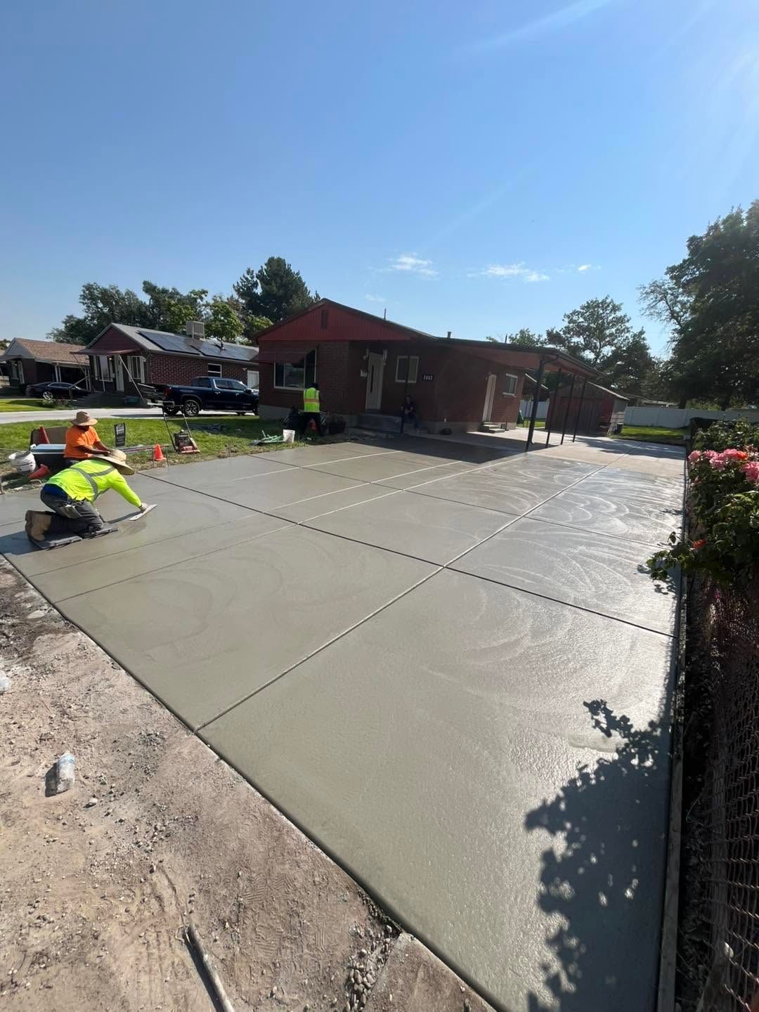 Workers smoothing a new concrete driveway beside a brick house on a sunny day