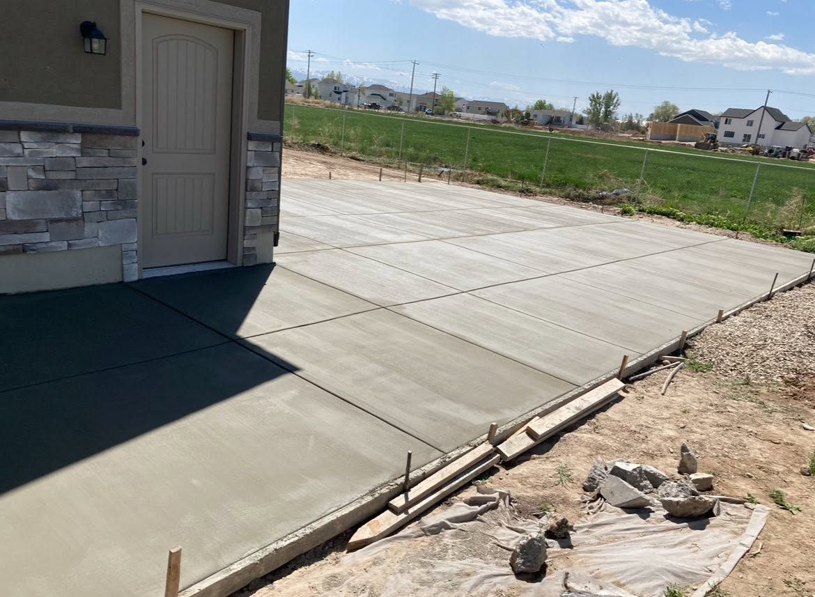 Fresh concrete driveway beside a house, bordered by gravel and open grassy land under a bright sky