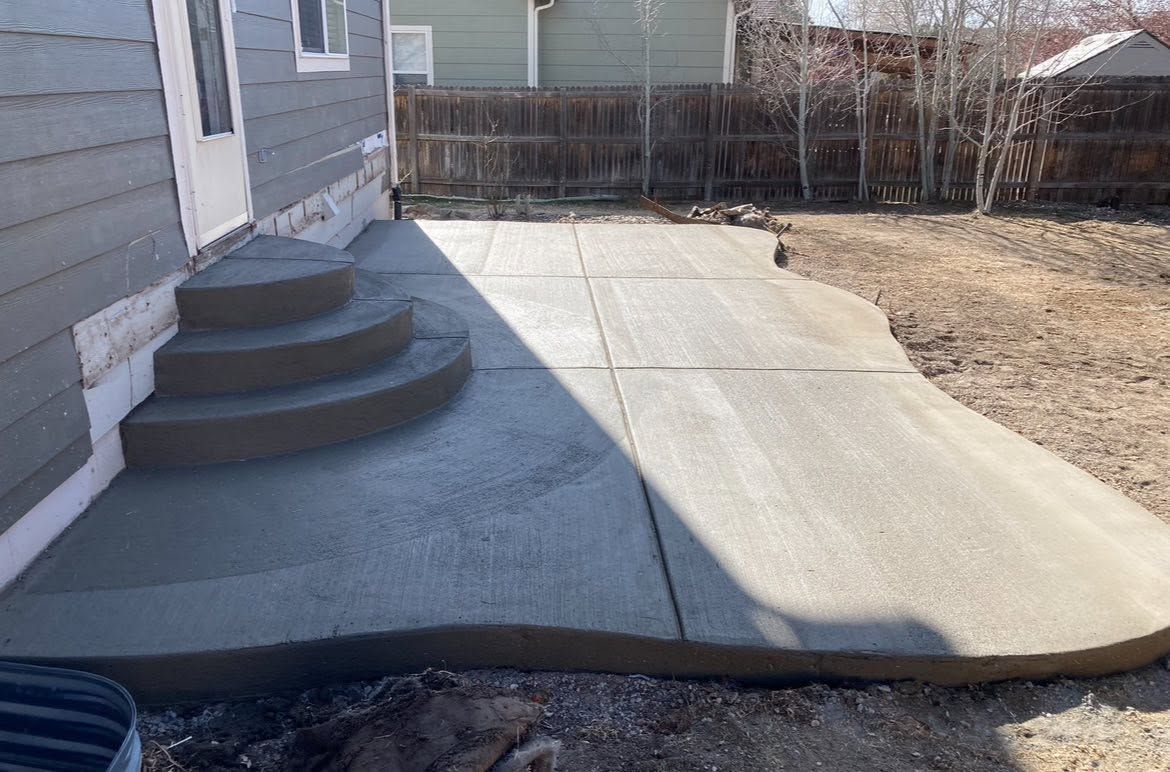 Concrete patio with curved steps beside a house, leading to a fenced backyard.