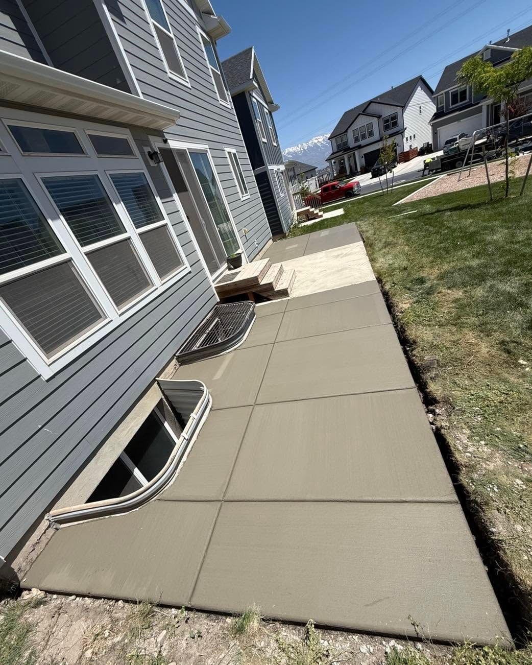House side walkway with concrete slabs beside gray siding and a patch of grass, leading to neighboring homes