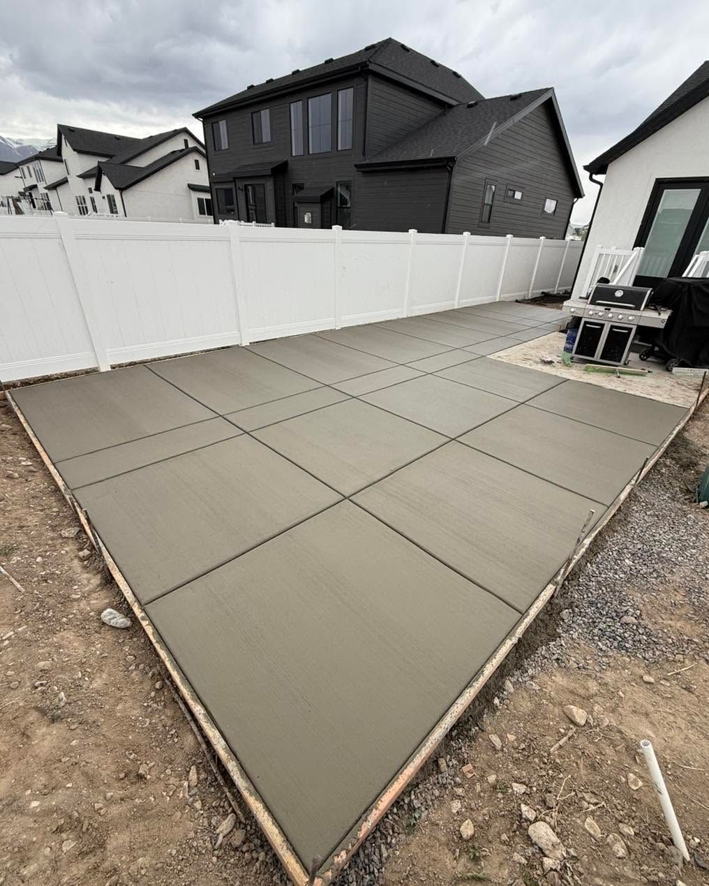 Fresh concrete patio slabs in a fenced backyard under construction, with neighboring houses in the background
