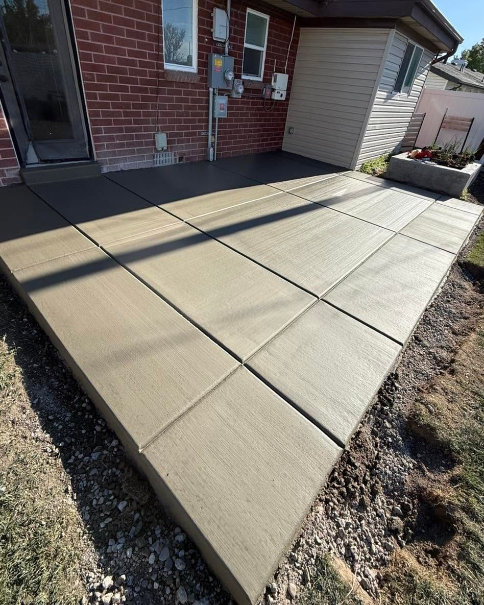 Concrete patio beside a brick house, with square slabs and gravel edging in bright sunlight.