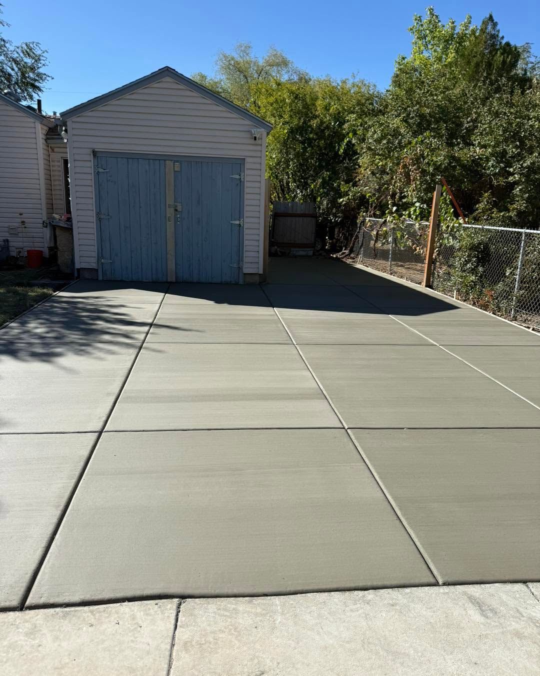 Concrete driveway leading to a small gray shed with blue doors, bordered by trees and a chain-link fence.