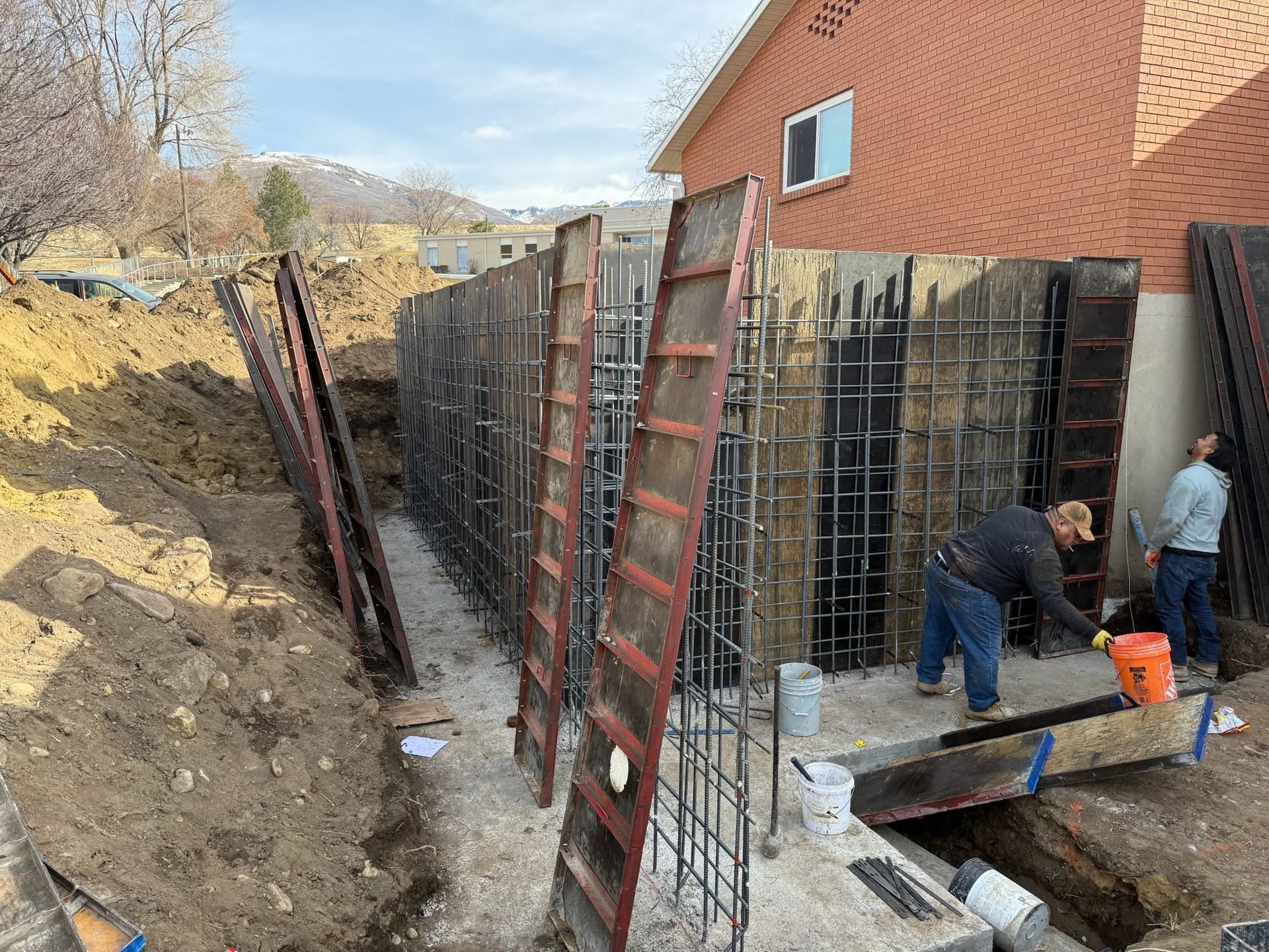 Construction crew working on a concrete foundation beside a brick house, with rebar forms and ladders.