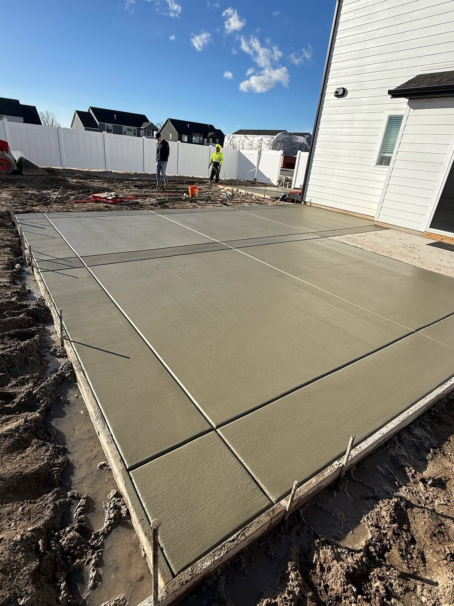 Fresh concrete slab poured at a construction site beside a white house, with workers in the background.