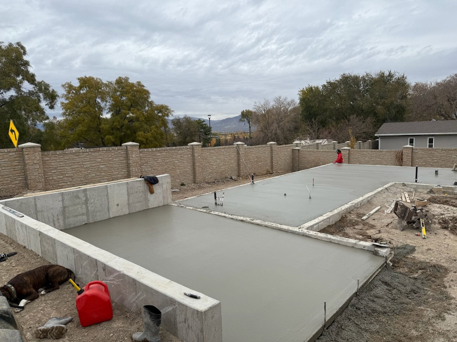 Concrete foundation under construction in a residential lot, with stone retaining walls and cloudy sky.