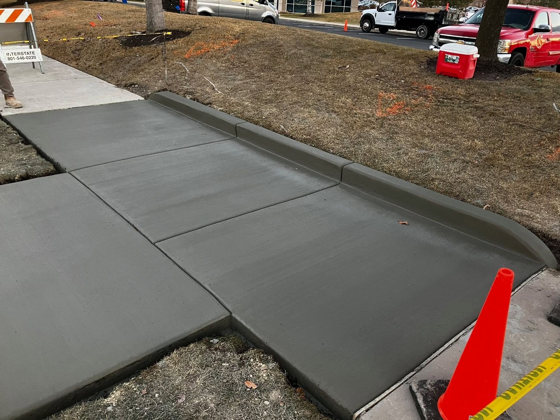 Fresh concrete sidewalk ramp beside a grassy area, marked by orange safety cones