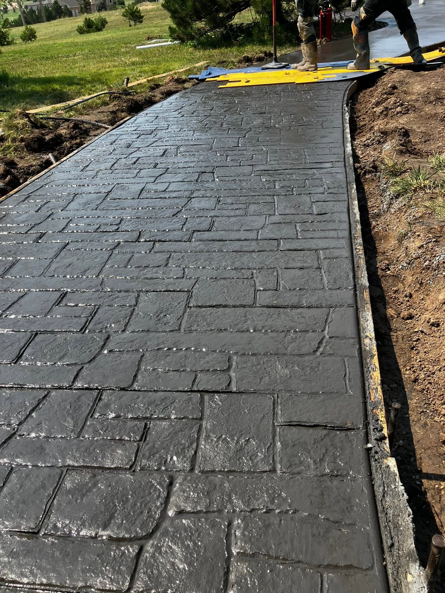 Freshly paved black walkway with workers and a yellow tool at the far end in a grassy outdoor area