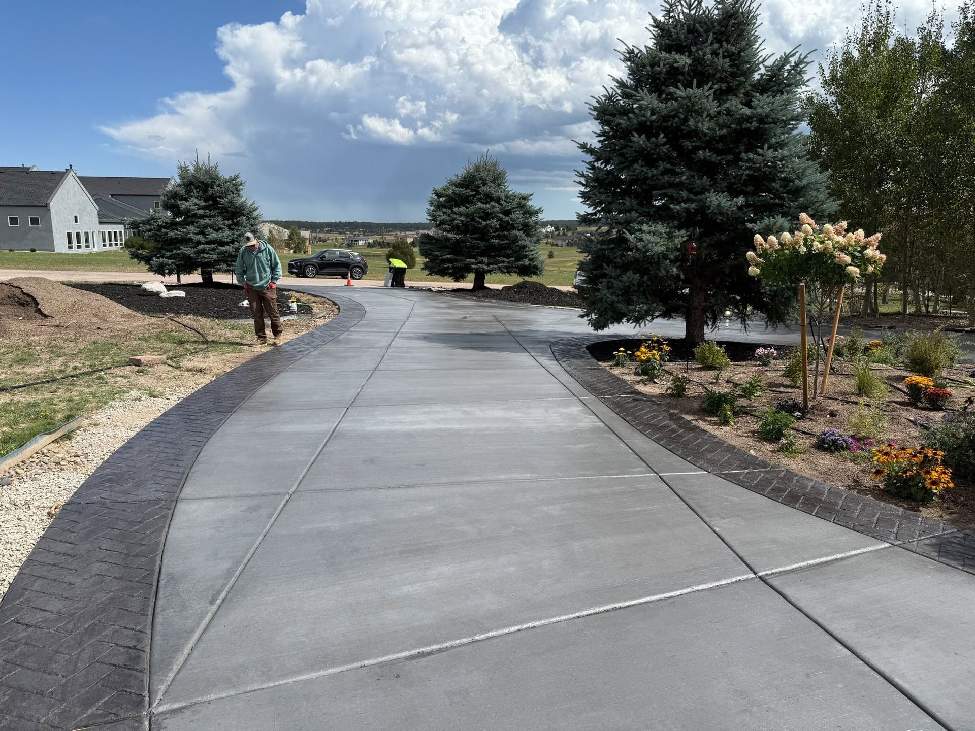 Curved concrete walkway through a landscaped neighborhood with trees, shrubs, and a worker in the distance