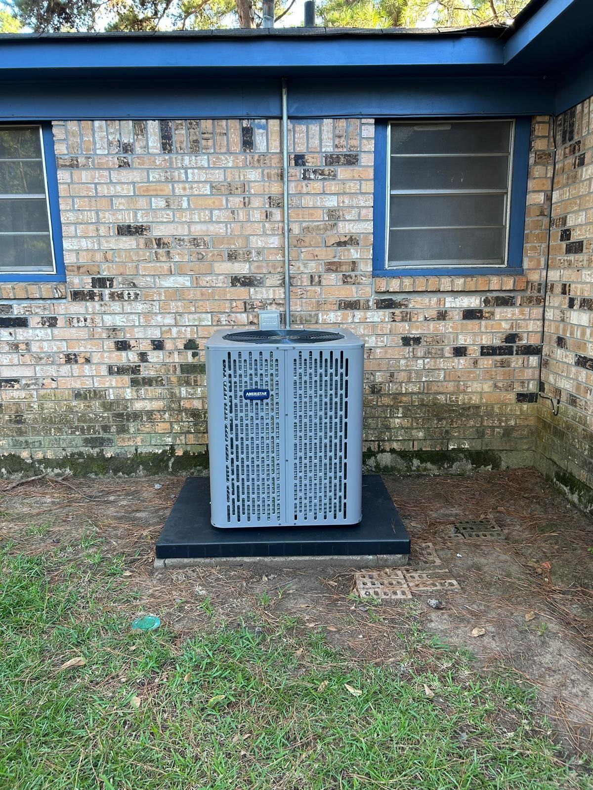 AC unit on a black platform against a brick wall with two windows and a blue trim.