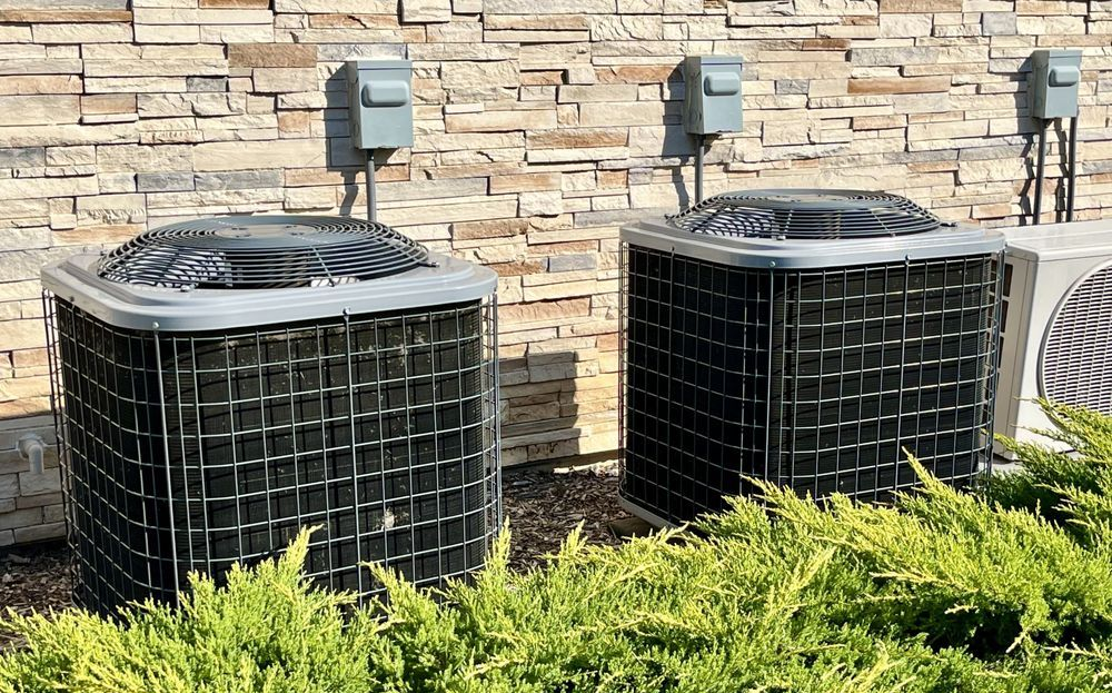 Two air conditioning units in front of a stone wall, with electrical boxes above, surrounded by green shrubs.