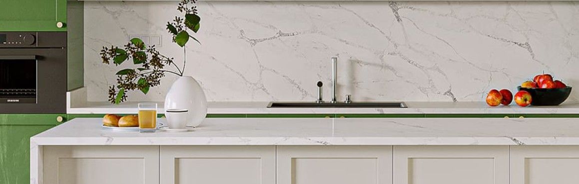 A kitchen with white countertops, green cabinets, and a marble backsplash. A vase with greenery and apples on the counter.