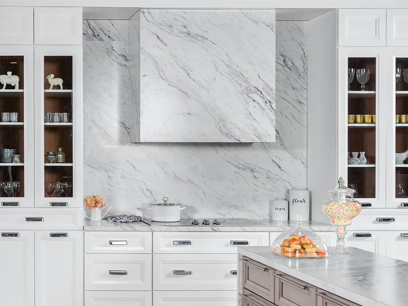 White kitchen with marble backsplash and range hood, glass-front cabinets, and an island.
