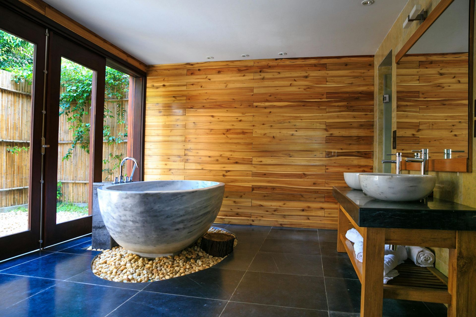 Bathroom with wooden walls, stone tub on pebbles, and black tiled floor, next to glass doors.