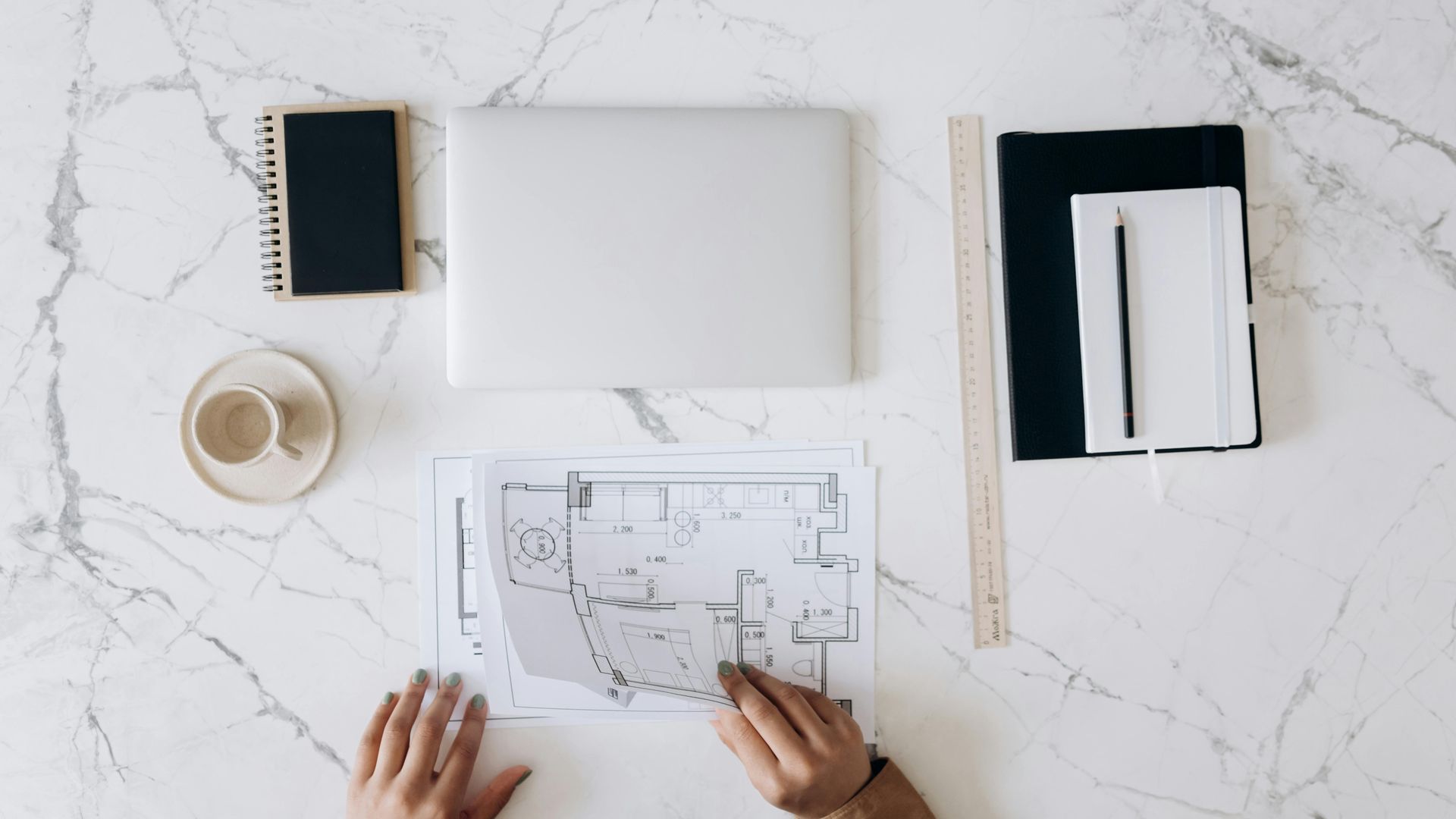 Hands unfolding a blueprint on a white marble surface, next to a laptop, notebooks, ruler, and a cup.