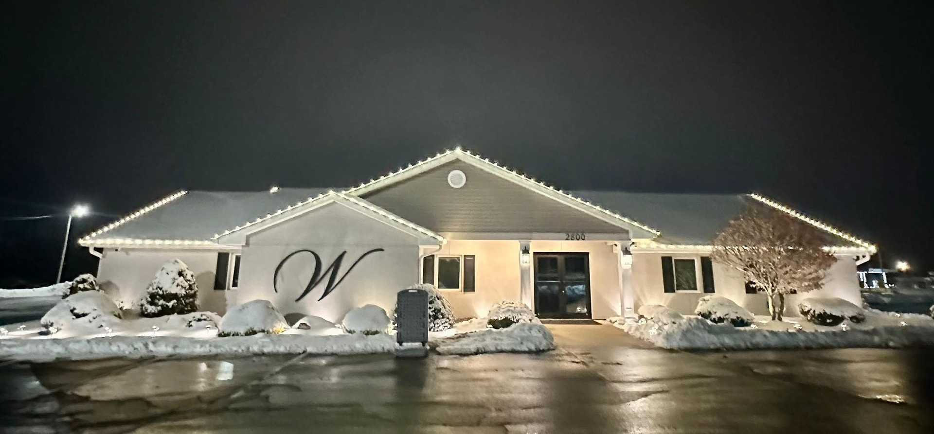 Snow-covered building at night with white holiday lights and wet pavement in front