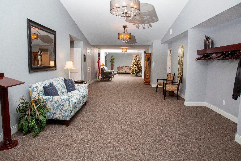 Carpeted hallway with floral sofa, potted plants, chairs, and chandeliers in a bright lobby area