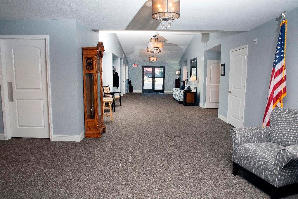 Wide hallway with gray carpet, light blue walls, wooden grandfather clock, and an American flag by an armchair.