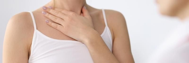 Persona che si tocca il collo e la gola, con indosso una canottiera bianca, su uno sfondo chiaro e uniforme.