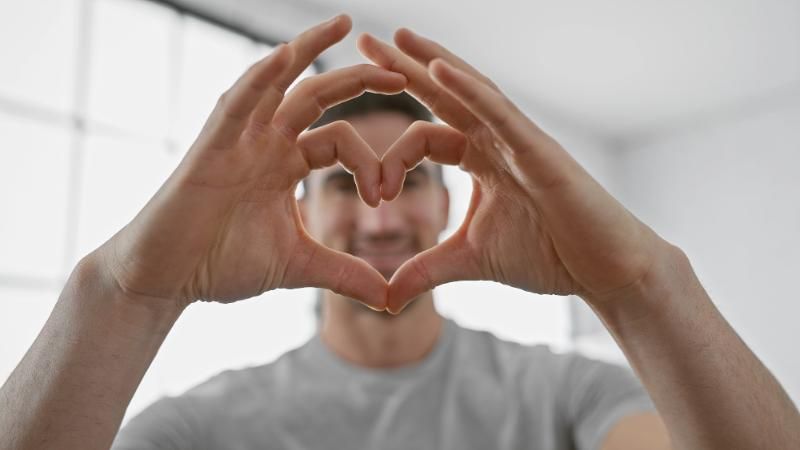 Mani che formano un cuore davanti a una persona sfocata in un ambiente interno.