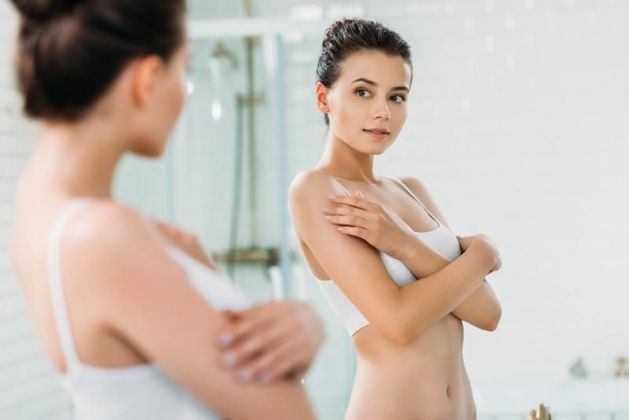 Una donna in biancheria intima bianca si osserva riflessa nello specchio del bagno.